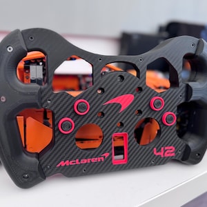 May include: A black and carbon fibre steering wheel with red detailing, displaying the McLaren logo and the number 42. The wheel has a complex design with multiple buttons and a textured grip. An orange toy car is visible in the background.