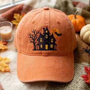 May include: An orange baseball cap featuring an embroidered design of a dark purple haunted house, a bare tree, a yellow full moon, and a black bat. The cap has a structured crown, a curved brim with stitching, and a button on the top center.