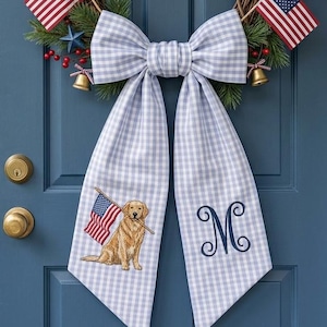 May include: A patriotic wreath with American flags, red, white, and blue stars, and berries hangs on a blue door. A large, light blue and white gingham bow with a golden retriever holding a flag and the letter "M" adorns the wreath. A hanging basket of flowers is also visible.