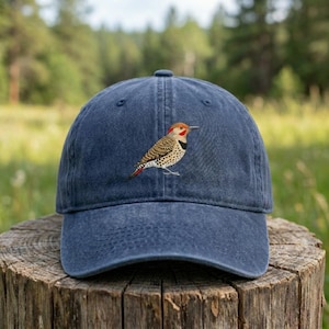May include: A navy blue baseball cap featuring an embroidered bird design. The bird has a red head, black markings, and a speckled pattern on its body. The cap is made of a soft, durable fabric.