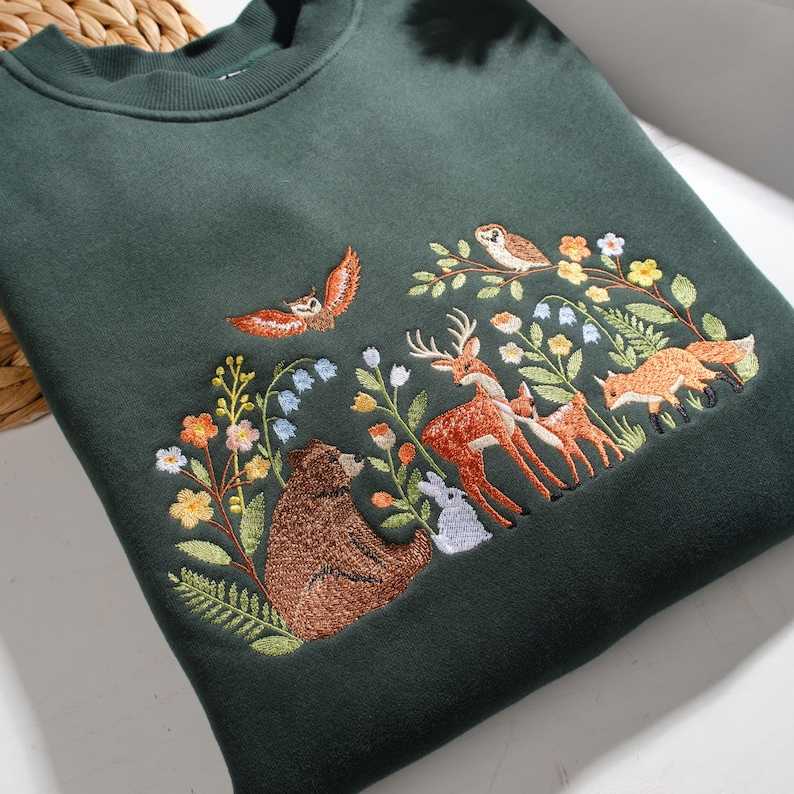 May include: A dark green crewneck sweatshirt featuring detailed embroidery of a forest scene. The design includes a brown bear, a deer with a fawn, a fox, a rabbit, and two owls surrounded by colorful wildflowers, green leaves, and ferns.