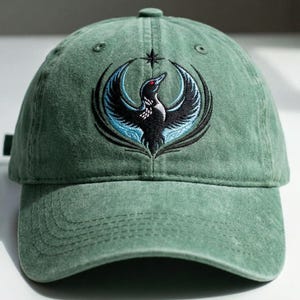 May include: Green baseball cap with an embroidered design of a black bird with blue and black wings, a red eye, and a star above it. The cap is made of a soft, textured fabric.