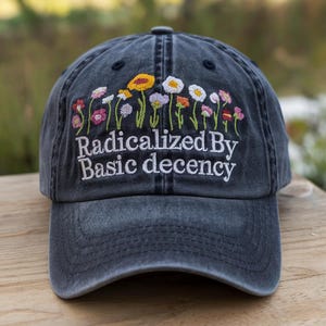 May include: A charcoal gray baseball cap featuring an embroidered design of colorful wildflowers across the top. Below the floral arrangement, the text "Radicalized By Basic decency" is stitched in white thread across the front panels.