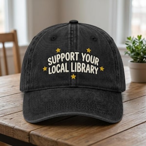 May include: Black baseball cap with the text "SUPPORT YOUR LOCAL LIBRARY" in white, accented with yellow stars. The cap is made of a worn-looking fabric and sits on a wooden surface.