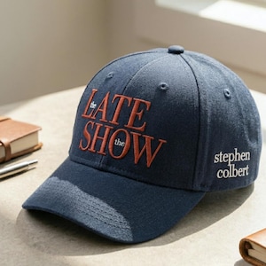 May include: Navy blue baseball cap featuring the words "the LATE SHOW the" in orange embroidery on the front. The name "stephen colbert" is embroidered in white on the side. A classic, adjustable hat.