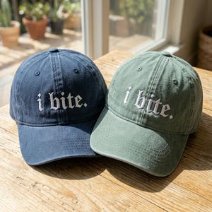 May include: Two baseball caps, one navy blue and one sage green, sit side-by-side. Each cap has the phrase "i bite." embroidered in white, gothic-style lettering. The caps are on a wooden surface, with natural light illuminating the scene.