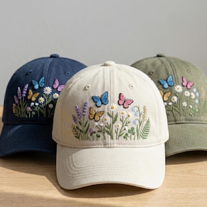 May include: Three baseball caps with embroidered butterfly and floral designs. The caps are in navy blue, cream, and olive green. The embroidery features butterflies in shades of blue, pink, and yellow, along with white daisies and lavender.