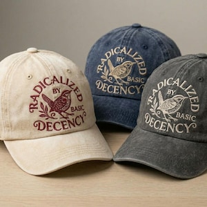 May include: Three baseball caps in beige, navy blue, and charcoal gray. Each cap features the embroidered text "RADICALIZED BY BASIC DECENCY" around a bird and branch design. The caps have a classic, adjustable fit.