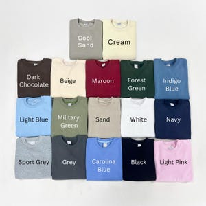 May include: A collection of seventeen crewneck sweatshirts arranged in a grid, featuring various solid colors labeled as Cool Sand, Cream, Dark Chocolate, Beige, Maroon, Forest Green, Indigo Blue, Light Blue, Military Green, Sand, White, Navy, Sport Grey, Grey, Carolina Blue, Black, and Light Pink.