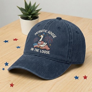 May include: A navy blue baseball cap with the text "Patriotic Goose on the Loose." The cap features a graphic of a goose wearing a hat and an American flag. Fireworks are also included in the design.