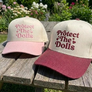 May include: Two baseball caps with the text "Protect The Dolls" embroidered on the front. One cap has a cream-colored crown and a pink brim, while the other has a cream-colored crown and a burgundy brim. The caps are sitting on a wooden surface.