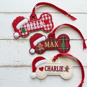 May include: Three bone-shaped felt ornaments with red ribbons. The top ornament is red and white gingham with the name BELLA, a heart, and a Santa hat. The middle is red plaid with MAX, a gingerbread man, and a Santa hat. The bottom is cream with CHARLIE, a heart, and a gingerbread man.