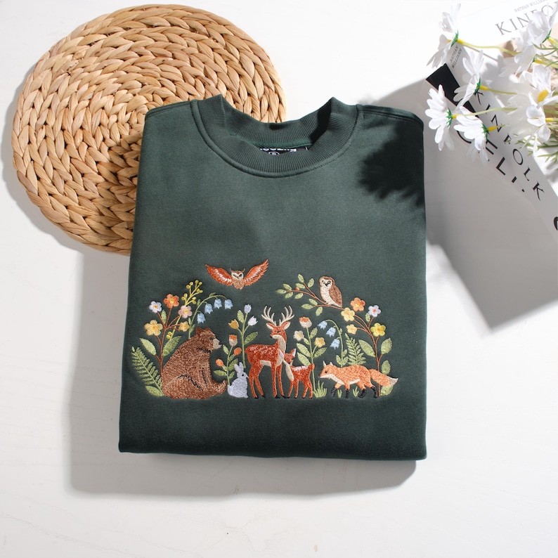 May include: A dark forest green crewneck sweatshirt featuring detailed embroidery of a woodland scene. The design includes a brown bear, a deer, a fawn, a fox, a rabbit, two owls, and various colorful wildflowers and green foliage.