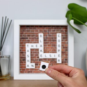 May include: A framed crossword puzzle with a brick background and white letters spelling out names. A small white square with a black dot is held in front of the puzzle. The frame is white.