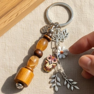 Puede incluir: Un llavero de madera con un anillo plateado y abalorios decorativos. Los abalorios incluyen un pequeño búho, una margarita blanca y hojas plateadas. La parte de madera tiene una serie de cuentas marrones claras y oscuras.