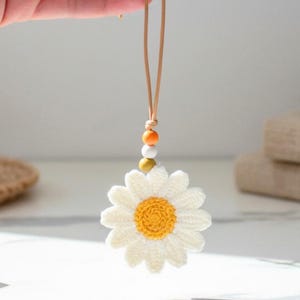 May include: A crocheted daisy ornament featuring white petals and a yellow center. The flower hangs from a thin tan cord accented with three small spherical beads in orange, white, and mustard yellow, creating a decorative hanging accessory.