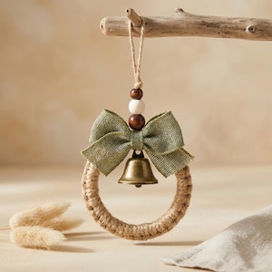 May include: A circular ornament wrapped in tan jute twine, featuring a sage green burlap bow, a metallic bronze-toned bell, and a vertical string of three wooden beads in dark brown, white, and dark brown, suspended by a thin jute loop.