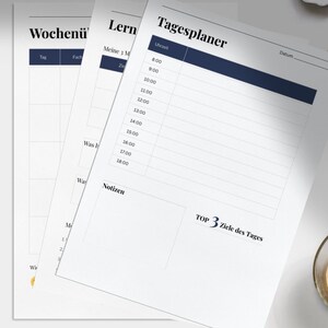 Op de afbeelding: Witte papieren planners met blauwe en zwarte tekst, waaronder een dagschema en een weekoverzicht. De afbeelding toont ook een kopje koffie en een pen op een wit oppervlak.