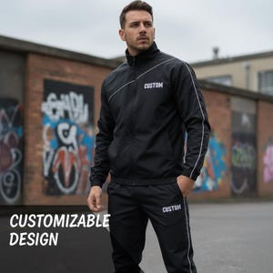 Op de afbeelding: Zwart trainingspak met witte biezen en het woord "CUSTOM" op de jas en broek. De jas heeft een hoge kraag en een diagonale witte streep. De tekst "CUSTOMIZABLE DESIGN" staat linksonder.