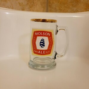 May include: A clear glass beer mug with a gold rim and a handle. The mug features a Molson Export Ale label with a red and white design and a blue ship illustration. The mug is sitting on a white surface.