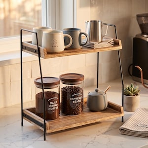 May include: A two-tiered wooden and metal shelf unit displaying coffee-related items. The top shelf holds mugs, a metal jug, and a folded cloth. The bottom shelf features glass jars with coffee beans and ground coffee, a small sugar bowl, and a succulent plant.