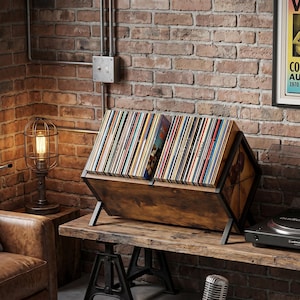 May include: A wooden record storage unit with a black metal frame, filled with vinyl records. The unit is on a rustic wooden table, with a vintage-style lamp and a record player visible in the background. The records are in various colours.