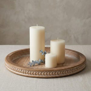 May include: Three cream-colored cylindrical pillar candles of varying heights arranged on a round wooden tray. The tray features a decorative beaded edge and a natural wood grain finish. A small sprig of dried eucalyptus leaves rests on the tray.