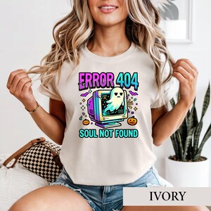 May include: An ivory t-shirt featuring a graphic of a vintage computer monitor with a ghost emerging from the screen. The text reads "ERROR 404" and "SOUL NOT FOUND" in bright blue and pink. Small orange pumpkins and purple bats surround the monitor.