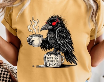 May include: Mustard yellow t-shirt featuring a black crow with red eyes holding a coffee cup with steam rising. The crow is perched on a coffee mug that reads "DON'T CAW ME UNTIL I'M BREWED".