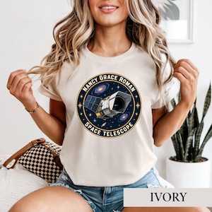 May include: An ivory short-sleeve t-shirt featuring a circular graphic with a dark blue background, gold stars, and a detailed illustration of the Nancy Grace Roman Space Telescope. The text reads NANCY GRACE ROMAN SPACE TELESCOPE.