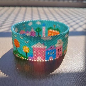 May include: A wide cuff bracelet crafted from small glass beads in a peyote stitch pattern. The design features a vibrant turquoise sky background with colourful houses in shades of pink, blue, and orange, along with green trees and white clouds.