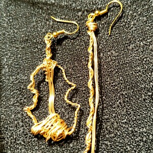 May include: A pair of handmade gold-colored wire earrings. One earring features a freeform design, while the other is a long, dangling style with a twisted wire element. The earrings are displayed on a dark textured surface.