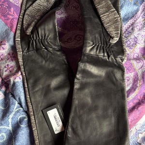 May include: A pair of black leather gloves with a gray knit accent on the back of the hand and along the side. The gloves are long, extending past the wrist. A product tag is attached to one glove.
