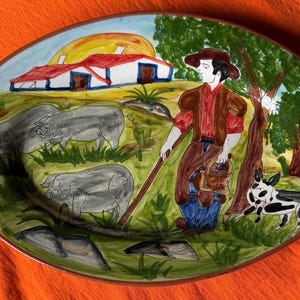 Puede incluir: Plato ovalado de cerámica con una escena pastoral pintada a mano. El diseño presenta un granjero, cerdos, un perro y una granja bajo un sol amarillo. El plato tiene un borde verde y está sobre un fondo naranja.