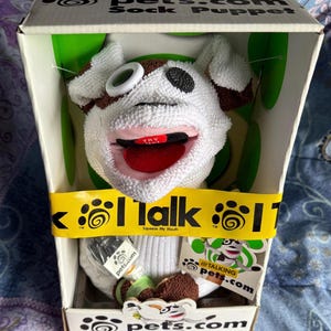 May include: A white and brown sock puppet with a large open mouth, red tongue, and a black and white eye, packaged in a box. The box has the text "pets.com Sock Puppet" and "I Talk". A tag reads "Talking Pets.com".