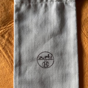 Pode incluir: Uma bolsa retangular cinza-claro com cordão, apresentando um padrão de espinha de peixe marrom-escuro e um logotipo circular marrom-escuro no centro. O logotipo retrata uma carruagem puxada por cavalos acima da letra H. Possui cordões marrons.