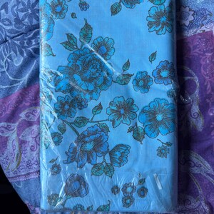 K&ouml;nnte beinhalten: Ein gefaltetes, hellblaues Bettw&auml;sche-Set mit Blumenmuster in Kunststoffverpackung. Das Blumenmuster zeigt gro&szlig;e blaue Blumen mit braunen und tealfarbenen Akzenten. Das Bettw&auml;sche-Set befindet sich auf einem gemusterten lila-blauen Hintergrund.