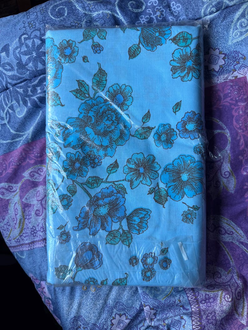 K&ouml;nnte beinhalten: Ein gefaltetes, hellblaues Bettw&auml;sche-Set mit Blumenmuster in Kunststoffverpackung. Das Blumenmuster zeigt gro&szlig;e blaue Blumen mit braunen und tealfarbenen Akzenten. Das Bettw&auml;sche-Set befindet sich auf einem gemusterten lila-blauen Hintergrund.
