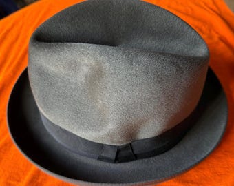 May include: A gray fedora hat with a dark gray brim and a black band. The hat has a classic, rounded crown and a slightly curved brim. The hat is made of a soft, felt-like material.