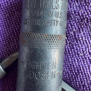 May include: A black Klein Tools 702 socket wrench with the text "MADE IN U.S.A." and "USE WITH SAFETY GOGGLES." The wrench has "TIGHTEN" and "LOOSEN" instructions. The wrench is on a purple surface.