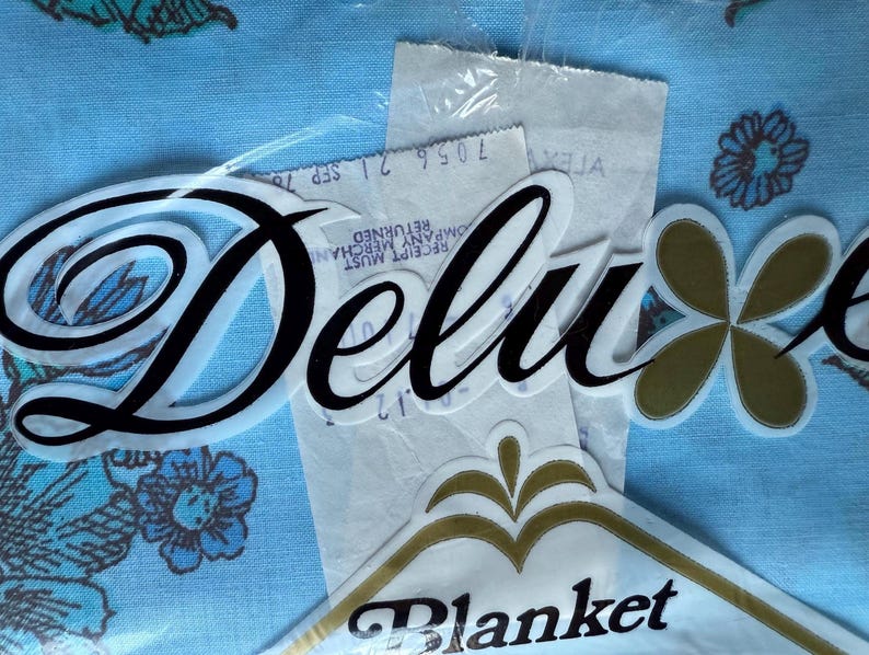 K&ouml;nnte beinhalten: Nahaufnahme einer Vintage-Deckenverpackung mit dem Wort "Deluxe" in Schreibschrift. Die Verpackung enth&auml;lt ein gold-wei&szlig;es Blumendesign und das Wort "Blanket". Der Hintergrund ist ein hellblauer Stoff mit floralen Mustern.