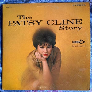 May include: Vintage vinyl record album cover for "The Patsy Cline Story." The cover features a portrait of Patsy Cline in a brown shirt, with the album title in bold black letters. The Decca logo and "Deluxe 2-Record Set" are also visible.