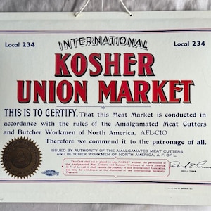 Op de afbeelding: Een ingelijst certificaat met de tekst "International Kosher Union Market" in rood en zwart. Het certificaat bevat tekst die de naleving van de regels van de markt certificeert en is uitgegeven door de Amalgamated Meat Cutters and Butcher Workmen of North America, AFL-CIO.