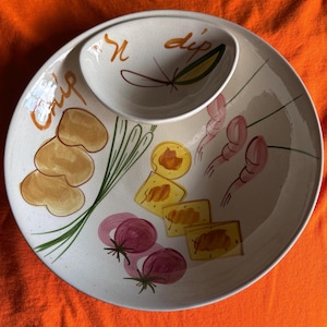 Op de afbeelding: Een witte keramische chip- en dipschaal met een ingebouwde dipschaal. De schaal is versierd met geschilderde illustraties van chips, crackers, uien en garnalen. De woorden "chip" en "dip" staan in oranje op de schaal.