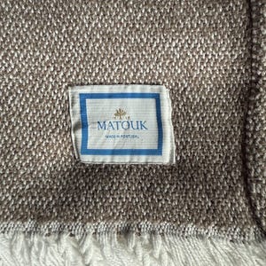 May include: A woven blanket with a textured brown and white pattern. The blanket has a white fringe along the bottom edge. A label with the brand name "Matouk" and "Made in Portugal" is sewn on the blanket.