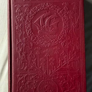 May include: A maroon-colored book titled "THE EBB-TIDE" with embossed floral and nautical designs. The cover features a sailing ship and decorative borders. The book is a biographical edition.