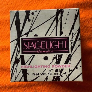 Puede incluir: Una caja de cosméticos cuadrada con un diseño abstracto en blanco y negro. La caja presenta el texto "STAGELIGHT Cosmetics" en un rectángulo rosa, con "HIGHLIGHTING POWDER" debajo. Peso neto 42,5 gramos.