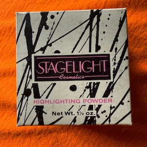 May include: A square box of Stagelight Cosmetics highlighting powder. The box has a white background with black paint splatter and lines. The text "STAGELIGHT Cosmetics" is in a pink rectangle. The text "HIGHLIGHTING POWDER" and "Net Wt. 1 1/2 oz." are also visible.