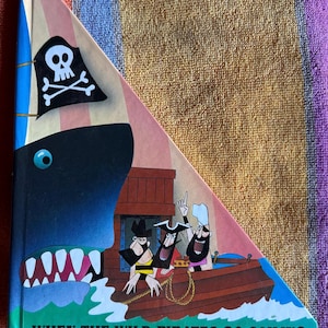 Może przedstawiać: Pop-up książka dla dzieci zatytułowana "When the Wild Pirates Go Sailing". Okładka przedstawia kreskówkową ilustrację piratów w łodzi, rekina i pirackiej flagi. Książka ma trójkątny kształt z kolorowym wzorem.