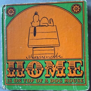 Pode incluir: Capa de livro vintage com Snoopy em sua casinha. A capa é laranja com uma borda verde e as palavras "HOME IS ON TOP OF A DOG HOUSE" em letras grandes e decorativas. Desenhos florais nos cantos.